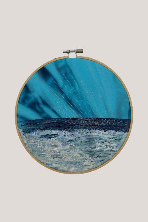 An embroidered seascape in a hoop, a dark stitched sea below a teal sky, pale beaded surf and shining thread catching a low sunset.